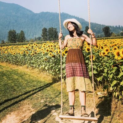 Isabella Wang stands on a rope swing in front of a field of sunflowers. She is smiling and wears a woven hat.
