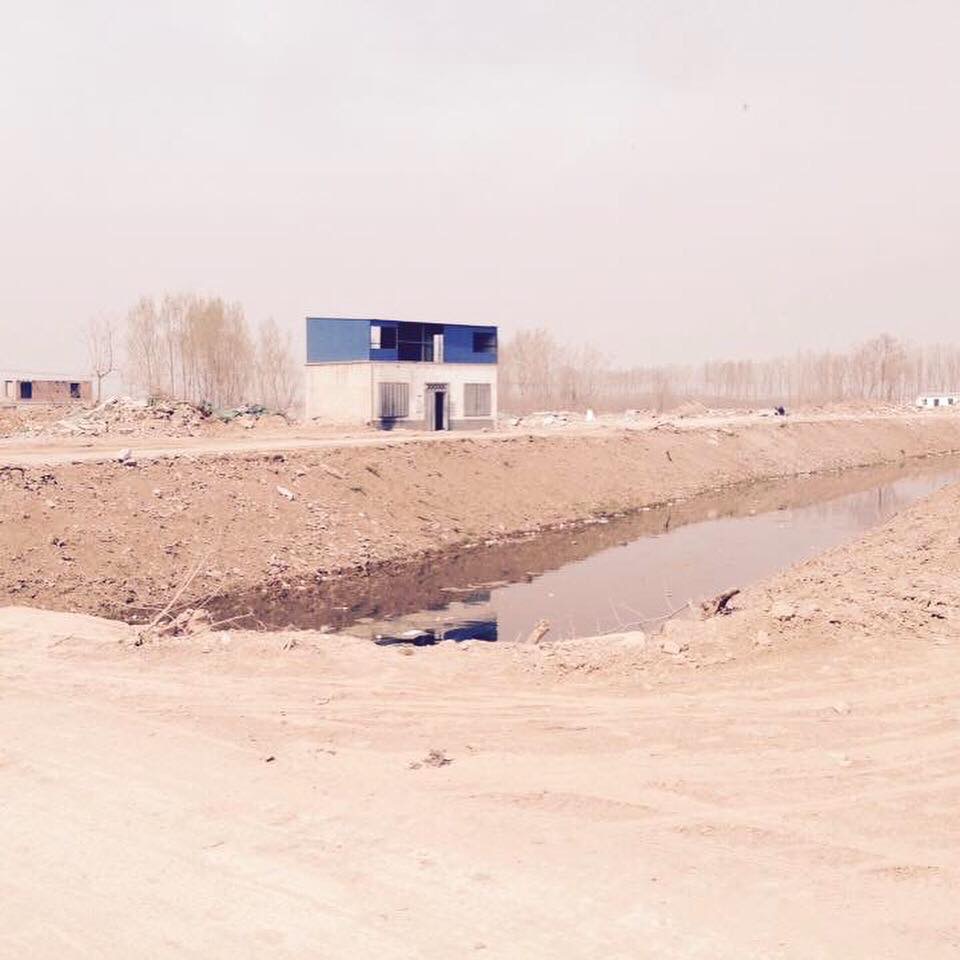 A dug out ditch full of water sits in front of a lone, two story, single room house. The structure is blue and white. Dirt and a few trees surround the structure.