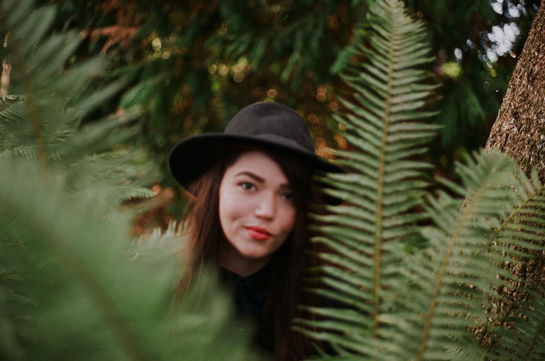 Author photo of Kayla MacInnis, wearing a black fedora, peering from behind a bush of fern leaves.