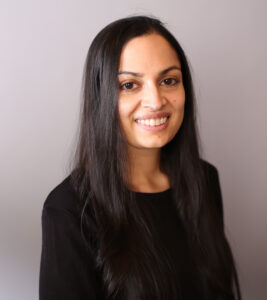 A South Asian woman smiles at the camera. She has long black hair, and is wearing a black full sleeved shirt.