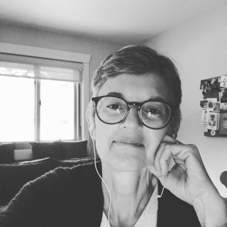 A short haired white woman smiles at the camera. She is wearing glasses and resting her hand on her cheek. The image is in black and white.