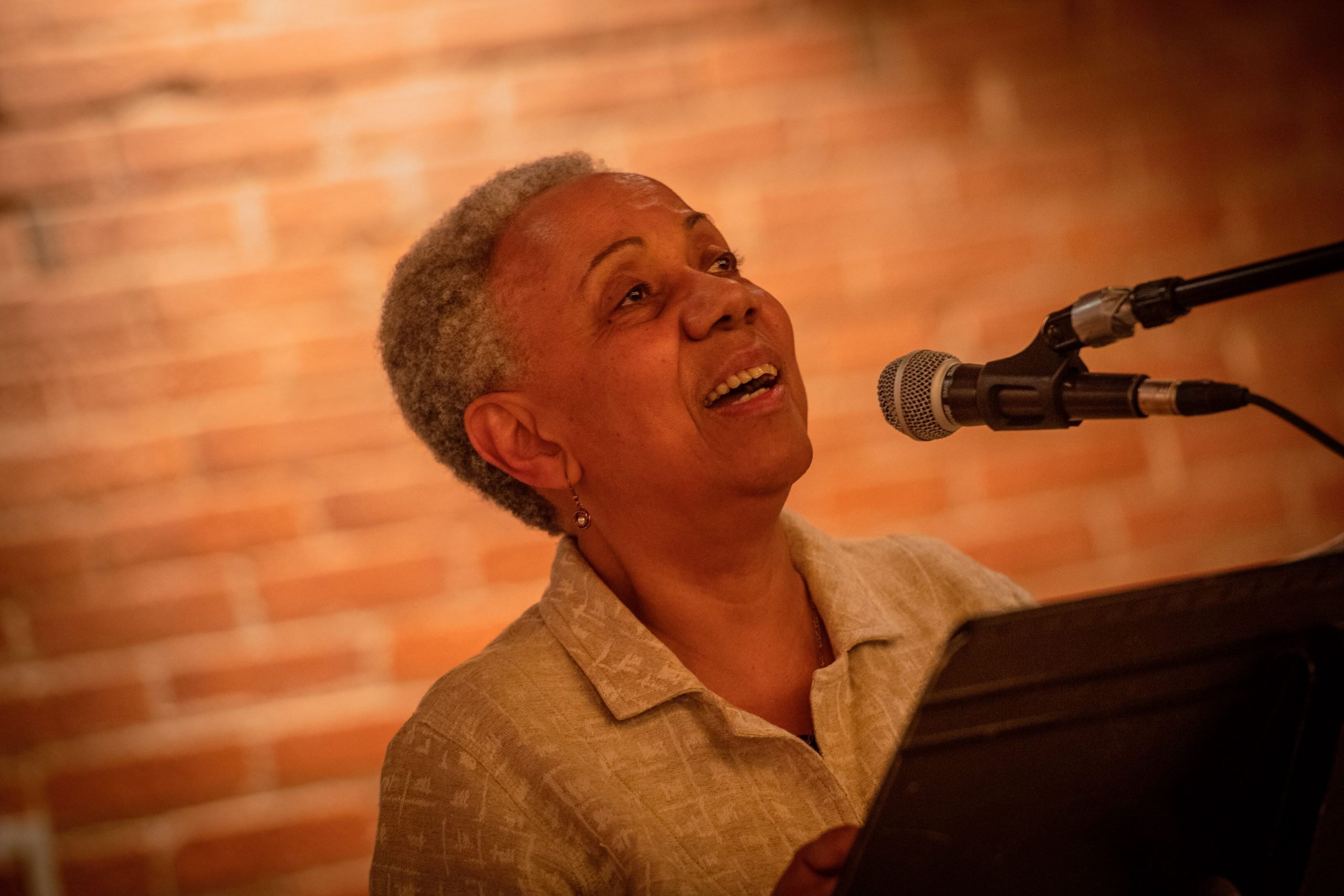 Lillian Allen speaks into a mic. She is standing in front of a brick wall.