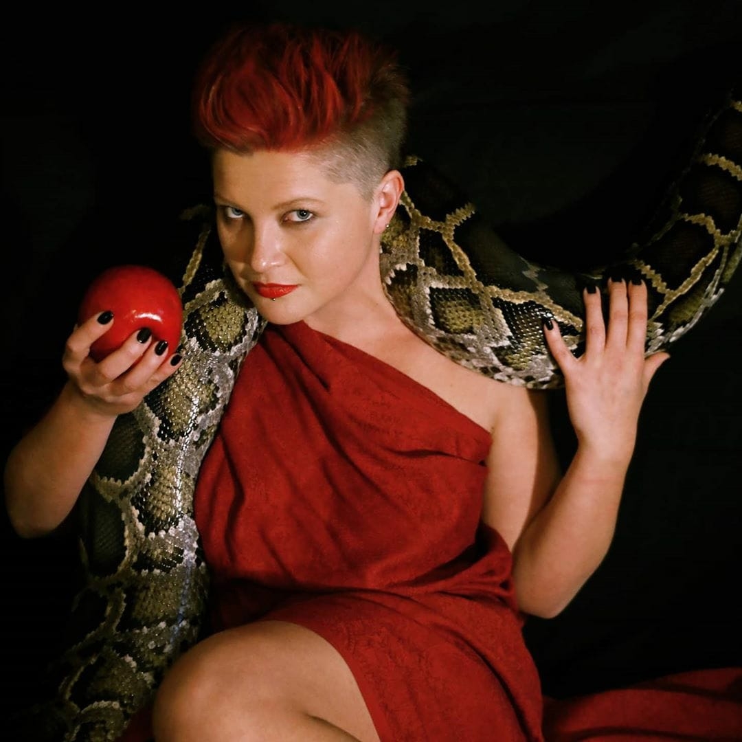 Vironika Wilde A person with red hair draped in a red cloth. They are holding an apple in one hand and a snake is wrapped around their shoulders.