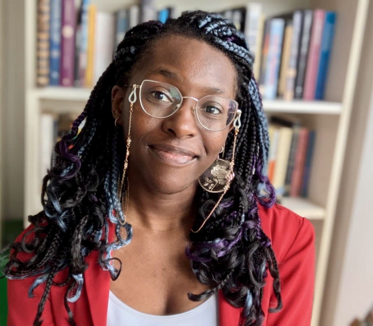 A headshot of Chimedum Ohaegbu. She's a young Black woman with braids, wearing glasses and a red jacket, smiling at the camera.