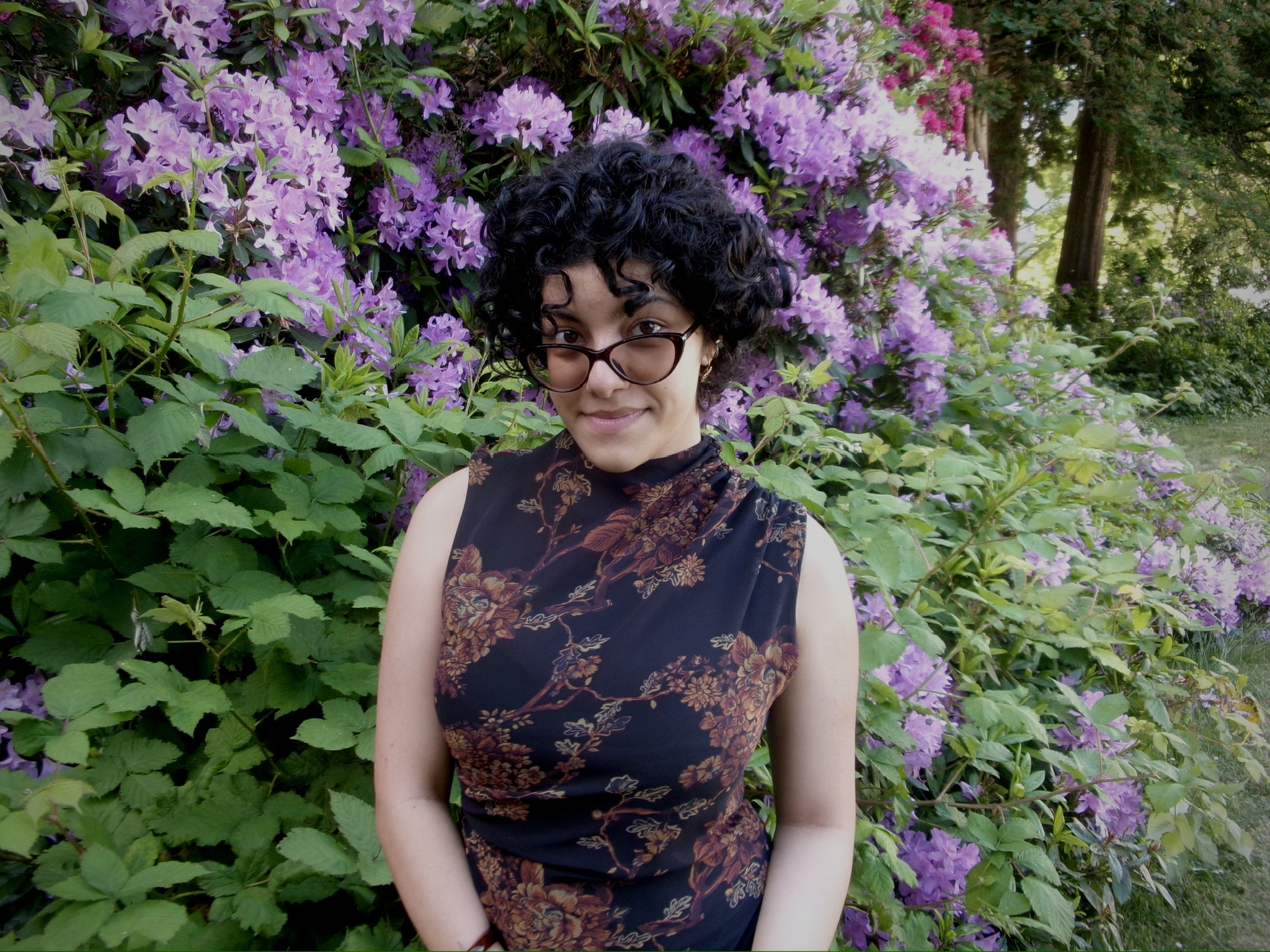 photo of Selin Berktas. She's a young Azerbaijani woman with curly dark hair and glasses, standing in front of a flowering bush.
